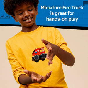 Young child wearing a yellow long-sleeved shirt smiling while holding a small red LEGO fire truck with black wheels against a blue and beige background with text reading "Miniature Fire Truck is great for hands-on play"
