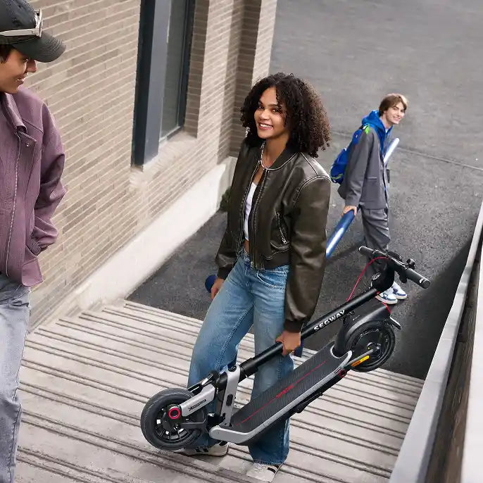 A woman with curly dark hair wearing an olive green leather jacket and blue jeans stands smiling while holding a black Segway Ninebot Kickscooter E3 Pro on a wooden deck, with two other people and a brick building visible in the background