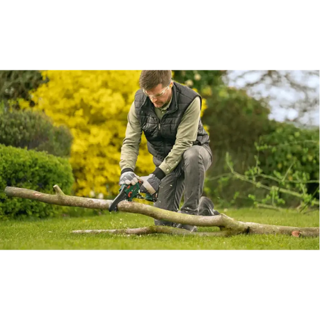 Man using Bosch Easy Chain 18V cordless chainsaw to cut wooden log on grass in garden with yellow flowering shrubs and green foliage in background