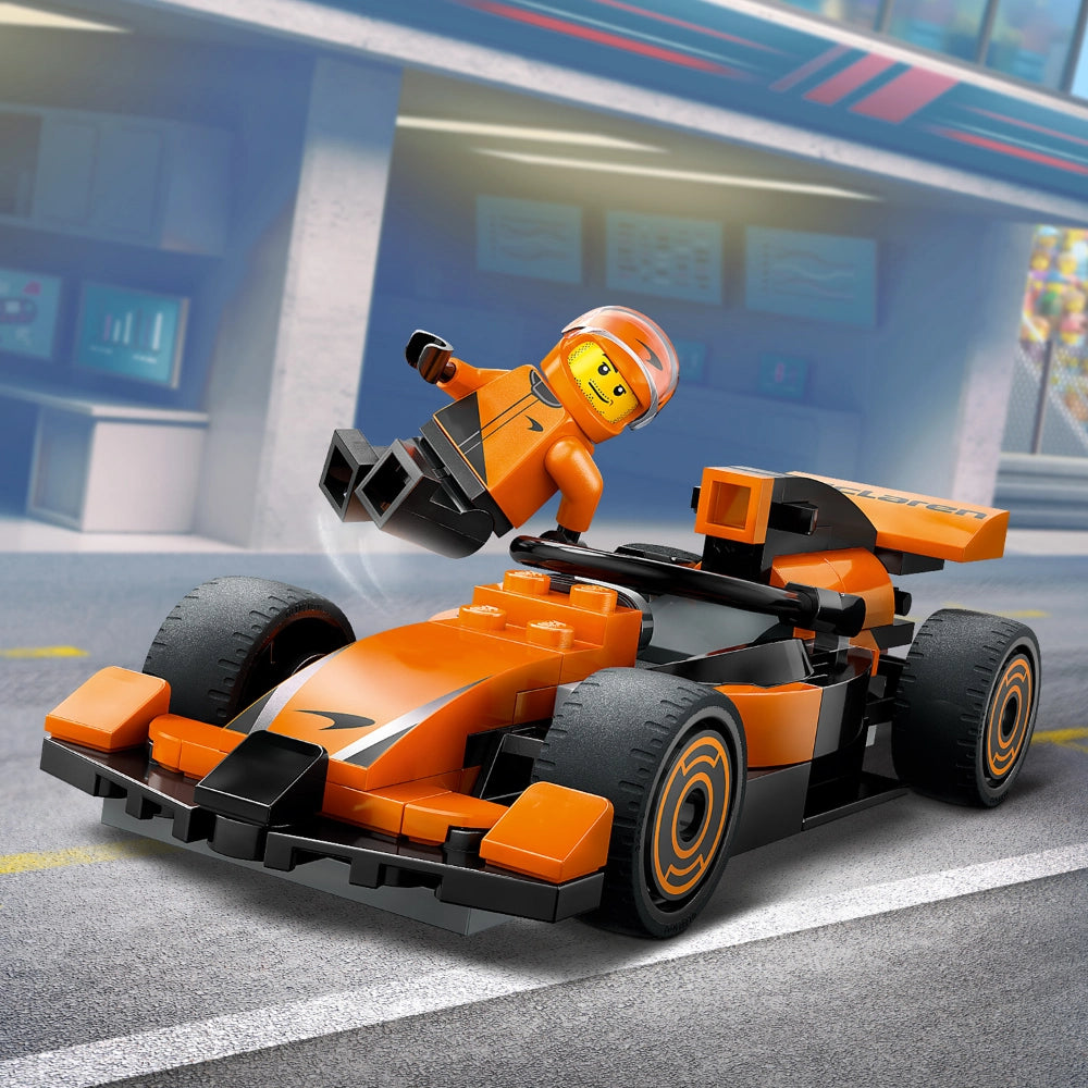 LEGO F1 driver minifigure in orange racing suit and helmet sitting in orange and black McLaren race car with grey wheels, displayed on grey race track with blurred pit garage background