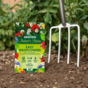 Unwins Nature's Haven Easy Wildflowers seed packet displayed on dark soil next to a white garden fork, with green foliage and flowering plants in the background, showing colorful wildflower illustrations and native British Isles flower varieties on the yellow-green packaging