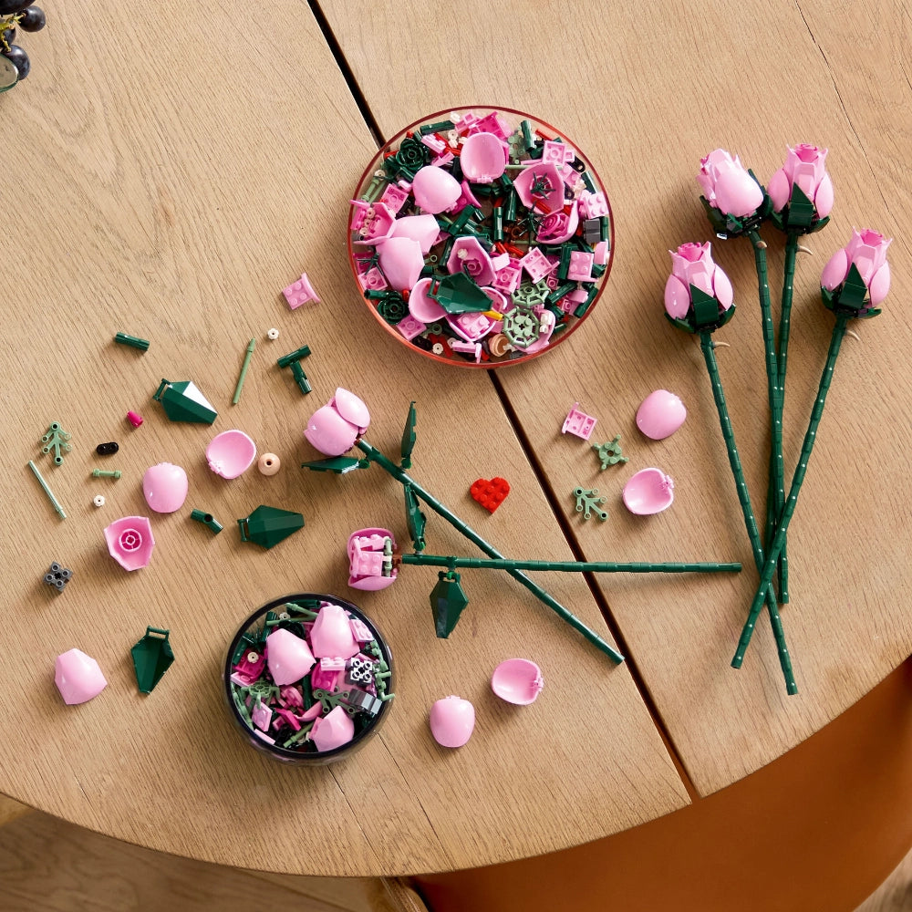 LEGO Bouquet of Pink Roses 10374 display showing assembled pink rose flowers with green stems, individual plastic components including pink petals, green leaves, and small accessories arranged on a wooden surface with two circular storage containers