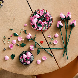 LEGO Bouquet of Pink Roses 10374 display showing assembled pink rose flowers with green stems, individual plastic components including pink petals, green leaves, and small accessories arranged on a wooden surface with two circular storage containers