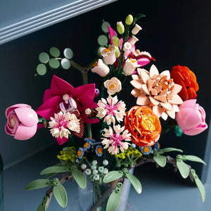 LEGO flower bouquet featuring pink roses, red and orange peonies, magenta orchids, cream daisies, yellow and white carnations with green stems and leaves in a clear glass vase against a dark background