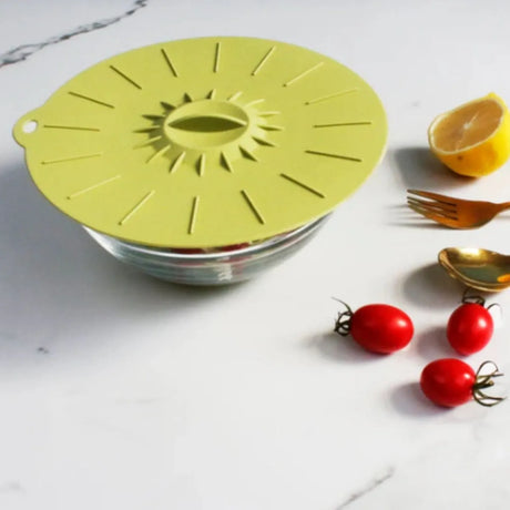 Yellow silicone suction lid on glass bowl with fresh red tomatoes, lemon wedge, and gold utensils displayed on white marble surface
