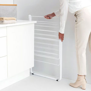 Woman in white clothing demonstrating the Brabantia HangOn Winged Clothes Airer, a white metal drying rack with multiple horizontal lines, positioned next to white kitchen cabinetry in a bright modern kitchen space