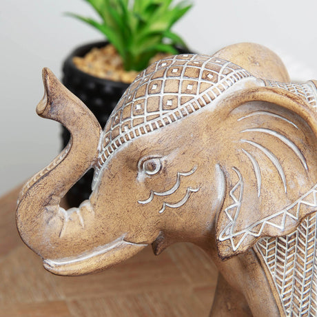 Tan stone elephant figurine with white embossed mirror detail patterns on body and ears, intricate carved designs, sitting pose with raised trunk, displayed on wooden surface next to small potted succulent plant