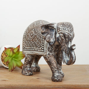 Bronze decorative elephant figurine with intricate Aztec geometric patterns on saddle blanket, featuring detailed trunk and tusks, displayed on wooden surface next to green succulent plant