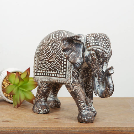 Bronze decorative elephant figurine with intricate Aztec geometric patterns on saddle blanket, featuring detailed trunk and tusks, displayed on wooden surface next to green succulent plant