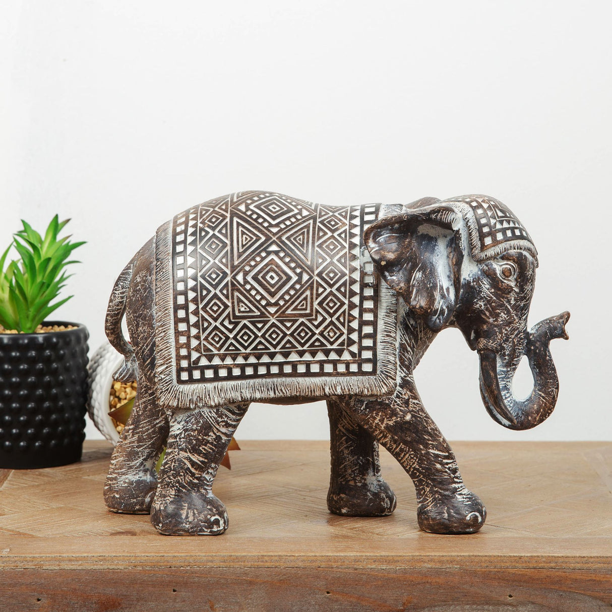 Bronze decorative elephant figurine with intricate Aztec geometric patterns and fringe detailing on saddle cloth, displayed on wooden surface alongside succulent plant in black pot
