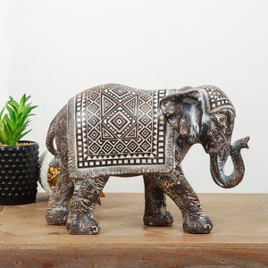 Bronze decorative elephant figurine with intricate Aztec geometric patterns and fringe detailing on saddle cloth, displayed on wooden surface alongside succulent plant in black pot