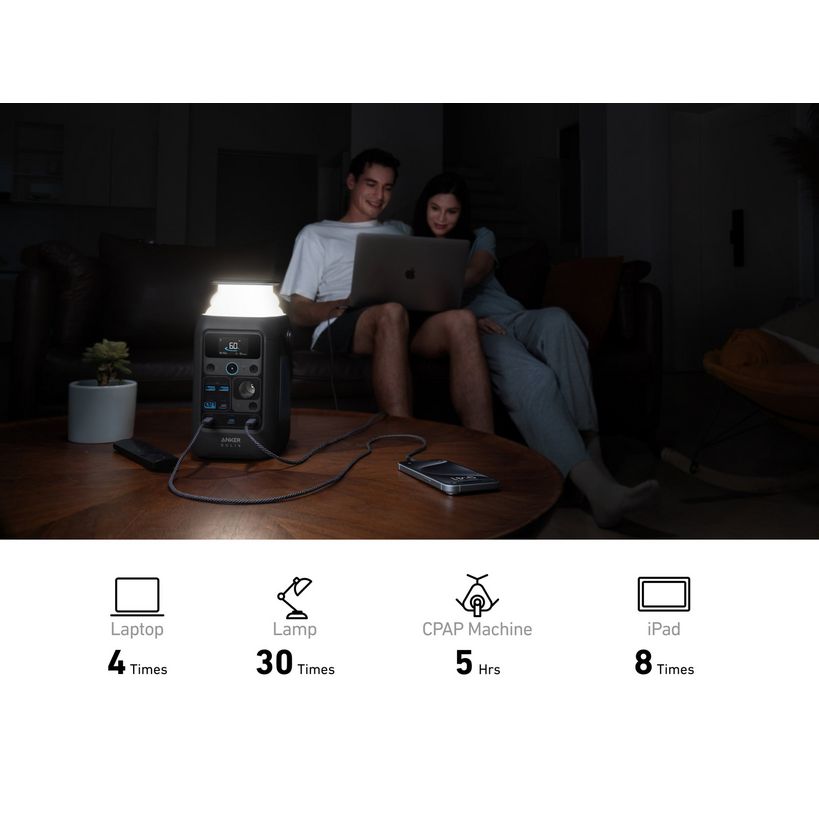 Anker Solix C300X portable power station displayed on wooden table powering a laptop, lamp, CPAP machine and iPad simultaneously in a dark living room with two people using the laptop in background
