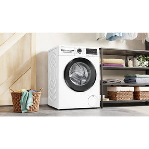 White Bosch Series 6 washing machine with black drum door in modern laundry room setting, surrounded by wicker laundry basket, wooden cabinetry, and storage shelves with folded towels and plants