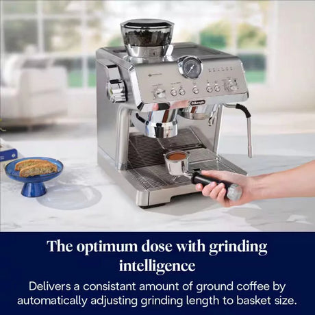 DeLonghi Magnifica stainless steel bean to cup espresso machine with integrated grinder dispensing espresso into a cup, featuring adjustable grind settings and control panel, displayed on a kitchen countertop with a blue plate of biscuits beside it