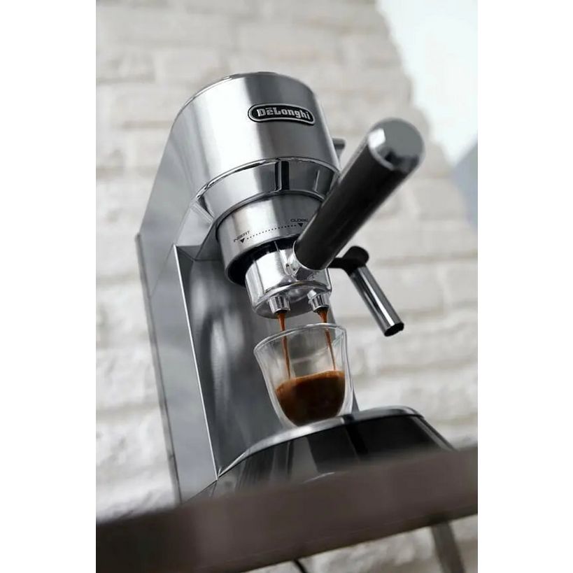 DeLonghi Dedica stainless steel pump espresso machine dispensing dark coffee into a clear glass cup, featuring chrome accents and black handle on a brick wall background