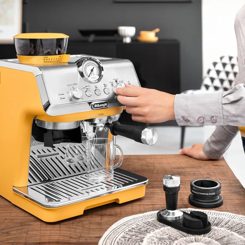 Person operating a yellow and stainless steel DeLonghi La Specialista Arte manual espresso machine on a wooden countertop, with espresso accessories and a black portafilter tamping guide visible in the foreground