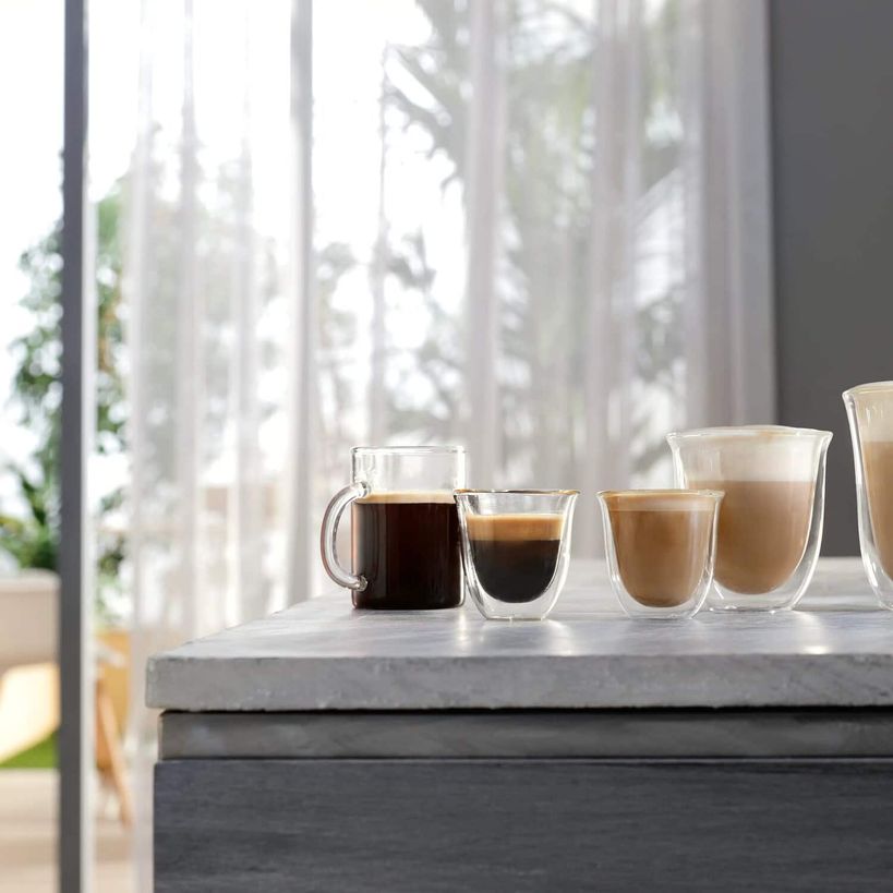 Various coffee drinks displayed in glass cups and a mug on a marble countertop showing espresso, americano, cappuccino and latte preparations with a bright window and sheer curtains in the background
