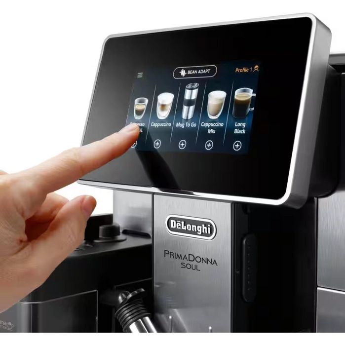 DeLonghi PrimaDonna Soul automatic coffee machine in metal black with touchscreen display showing coffee drink options including espresso, cappuccino, and long black, with a hand selecting a beverage profile