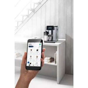 Hand holding smartphone displaying DeLonghi coffee machine app with various espresso and cappuccino options, positioned in front of the DeLonghi PrimaDonna Soul automatic coffee machine in metal black finish sitting on a white shelf in a minimalist white interior with stairs visible in background