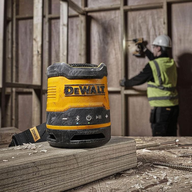 DeWalt DCR009 compact Bluetooth speaker in yellow and black on wooden workbench with construction worker in hard hat and safety vest using power tools in blurred background, industrial warehouse setting