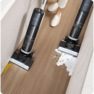 Overhead view of Dreame H14 Pro wet and dry vacuum cleaner demonstrating cleaning capabilities on wooden floor with spilled colorful items and liquid, showing two black cylindrical units with white cleaning pads and a person wearing white slippers