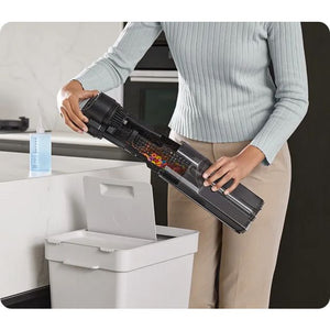Woman in light gray ribbed sweater and beige pants holding black Dreame H14 Pro wet and dry vacuum cleaner over white waste bin in modern home office setting