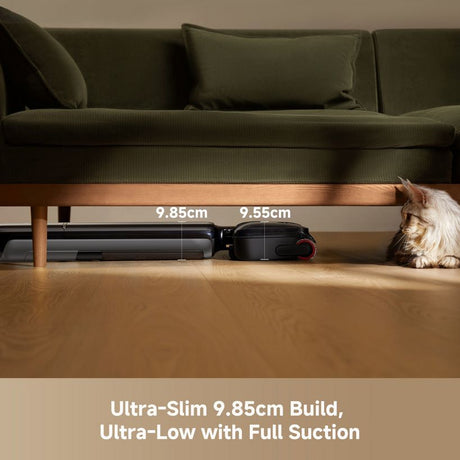 Dreame T12 Pro cordless vacuum cleaner demonstrating ultra-slim 9.85cm build height fitting under wooden sofa frame with green cushions, showing black vacuum unit next to measurement markers with cat beside it on light wood flooring