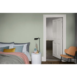 Bedroom painted in soft sage green Dulux Tranquil Dawn with bed featuring grey and blue pillows, pink throw, and yellow accent cushion, white bedside table, black floor lamp, tan leather chair, and white four-panel closet doors on dark wood flooring