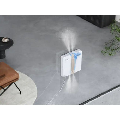 Ecovacs Winbot Mini beige and white robotic window vacuum actively cleaning a glass window with water spray visible, mounted on exterior glass with power cord, shown in modern outdoor patio setting with concrete flooring and potted plants