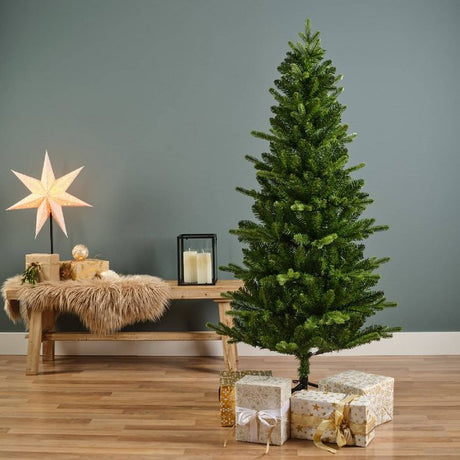Everlands Killington Fir Christmas Tree 6ft displayed in a modern living room setting with wooden bench, cream and gold wrapped presents, illuminated star decoration, candles in black lantern, and grey wall background