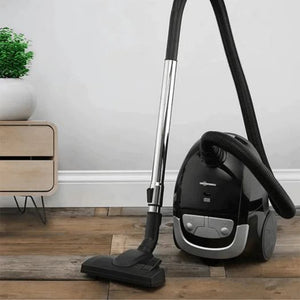 Black and silver bagged vacuum cleaner with hose attachment on wooden floor next to wooden nightstand with potted plant, demonstrating product in home setting