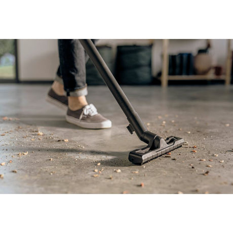 Person using black Kärcher WD4 V20 wet and dry vacuum with floor brush attachment to clean concrete floor covered in debris, wearing casual sneakers and dark pants indoors
