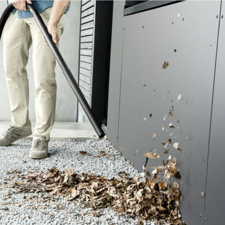 Man in cream pants using Kärcher WD 2 Plus wet and dry vacuum cleaner to suction up brown debris and gravel from concrete floor next to gray cabinet in indoor garage setting