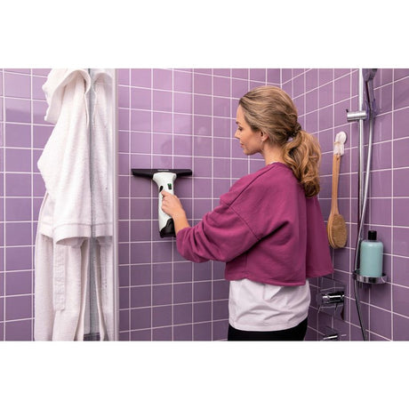 Woman using Karcher WV2 Plus window vacuum cleaner on purple tiled bathroom wall, with white towel and cleaning supplies visible
