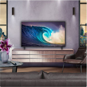 Modern living room displaying LG NANO81 55-inch 4K UHD Smart TV mounted on purple accent wall above light wood media console, showing turquoise wave imagery on screen, with dark leather sofa, brown accent chair, small round table with pink flowers in white vase, and pendant lighting