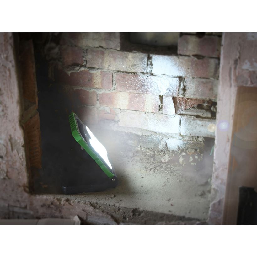 Lighthouse rechargeable worklight with 2000 lumens illuminating a brick wall in a dark basement or construction space, showing the bright white LED beam with green accent stripe on the black housing