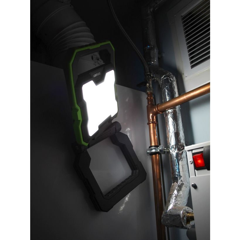 Lighthouse rechargeable worklight with 2000 lumens featuring bright white LED panel in green and black casing, mounted on wall next to copper pipe and ventilation unit in dark workshop setting