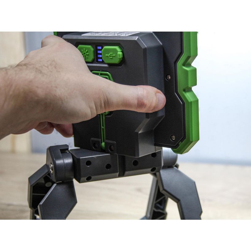 Hand holding green and black rechargeable work light with LED display and tripod stand base on wooden surface
