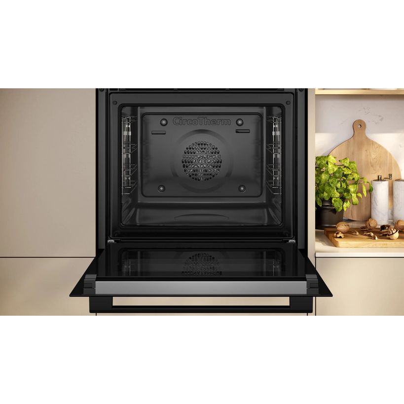 Black Neff N30 single built-in pyrolytic oven with open door displaying stainless steel interior and cooling fan, integrated into beige kitchen cabinetry with wooden chopping board and potted herb visible on shelf to the right