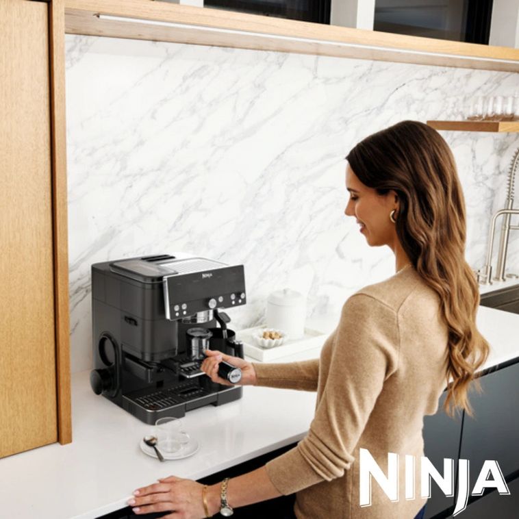 Woman in beige cardigan operating black Ninja Luxe Café Premier Series coffee machine on white marble backsplash countertop in modern kitchen with wooden cabinetry