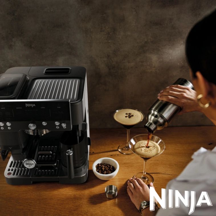 Ninja Luxe Café Premier Series black coffee machine on wooden counter with person pouring espresso-based drink into martini glass, coffee beans in white bowl, and Ninja branding visible