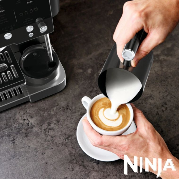Barista pouring steamed milk from a black pitcher into a white cup of espresso with latte art, next to a Ninja Luxe Café Premier Series black coffee machine on a dark grey surface
