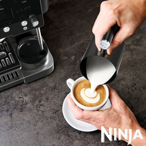 Barista pouring steamed milk from a black pitcher into a white cup of espresso with latte art, next to a Ninja Luxe Café Premier Series black coffee machine on a dark grey surface