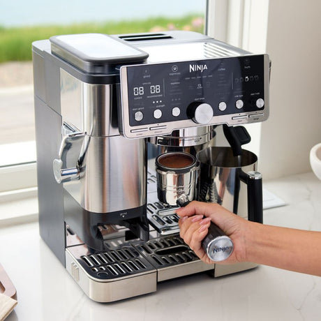 Ninja Luxe Café Pro Series stainless steel espresso machine with black control panel, digital display showing 08:08, dispensing rich brown espresso into a metal cup, hand holding Ninja-branded portafilter, window view in background