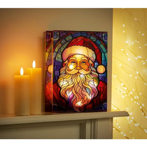 Battery operated LED Santa stained glass table top decoration with colorful red and gold design, displayed on white shelf with lit candles and warm golden lighting in background