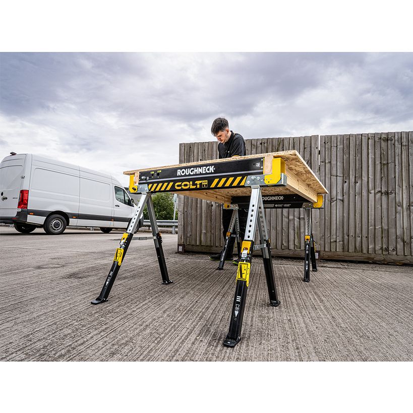 Roughneck Colt yellow and black folding sawhorse with worker on platform in industrial yard next to white van and wooden fence