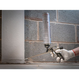 Worker in white gloves holding Roughneck expanding foam gun with cylindrical cartridge against concrete block wall during application