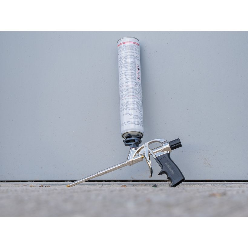 Roughneck expanding foam gun with white cartridge cylinder and black trigger handle, laid horizontally against gray background and concrete surface
