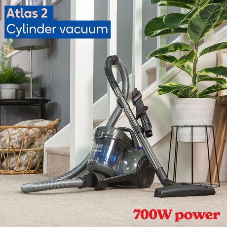 Russell Hobbs Atlas 2 cylinder vacuum cleaner with 700W power in dark blue and black, displayed on beige carpet in a modern living room with white pillars, potted plants, and wooden furniture