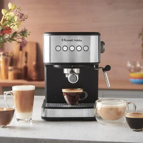 Russell Hobbs Everyday Espresso black and stainless steel coffee maker with five control buttons dispensing espresso into glass cup, surrounded by various coffee beverages on white countertop with blurred kitchen background